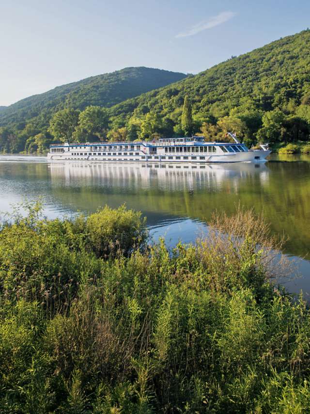 River cruise ship sailing along a calm river surrounded by forested hills.